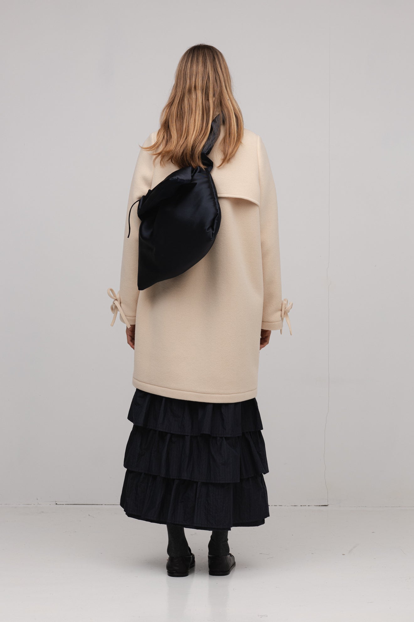 Person wearing a beige coat and black ruffled skirt with a black bag on a white background