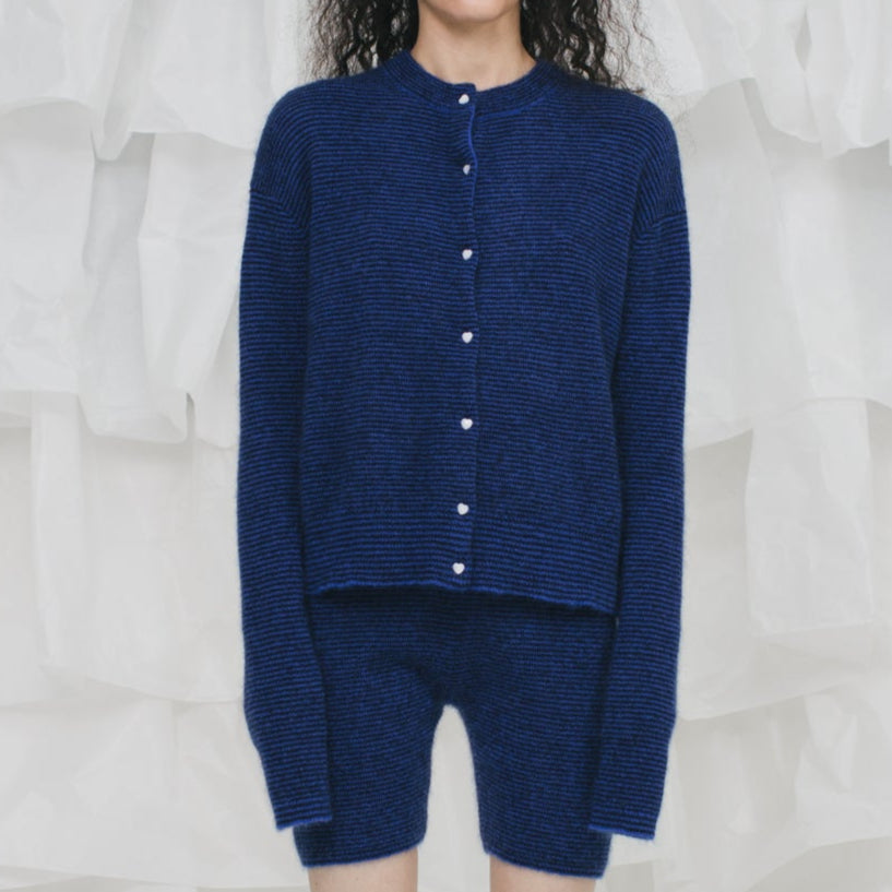 Person wearing a blue knitted cardigan and shorts against a white curtain background